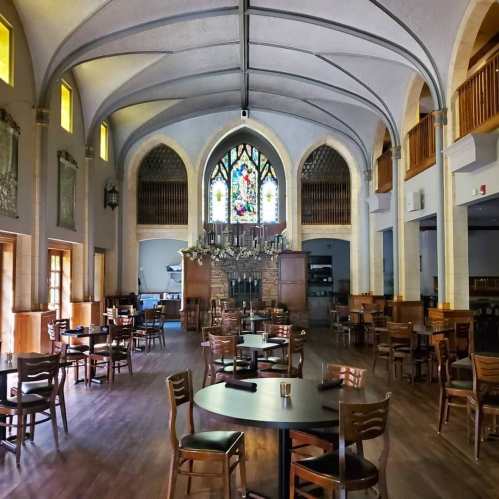 A spacious restaurant with wooden tables and chairs, featuring a stained glass window and high arched ceilings.
