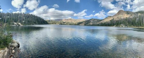A serene lake surrounded by mountains and trees, under a blue sky with fluffy clouds.