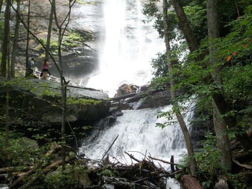 A serene waterfall cascades over rocks, surrounded by lush greenery and trees, with two people in the background.