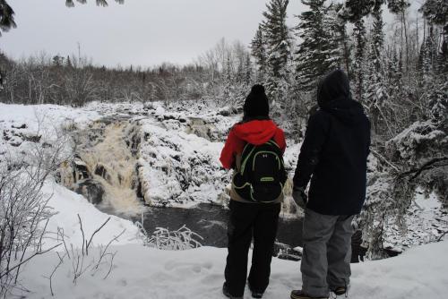 Two people stand by a snowy riverbank, gazing at a waterfall surrounded by trees in winter.