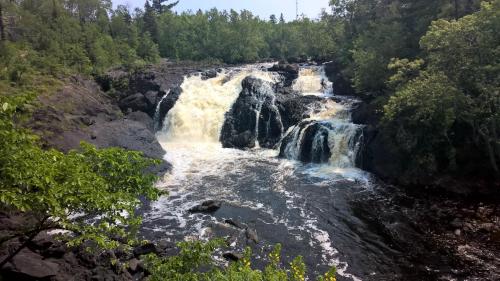 A scenic waterfall cascading over rocky terrain, surrounded by lush green trees and a clear blue sky.
