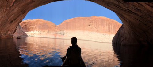A silhouette of a person in a kayak inside a cave, with red rock formations and a blue sky visible in the background.