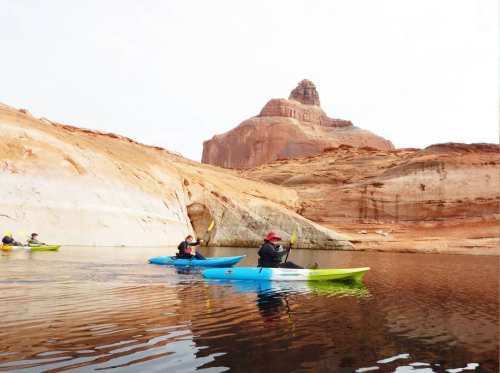 Two kayakers paddle on calm water near rocky cliffs, with a prominent rock formation in the background.