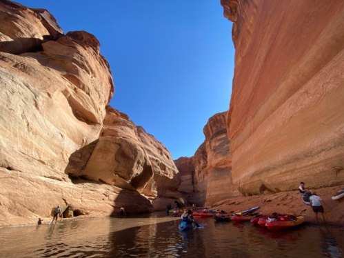A serene canyon with towering red rock walls, people kayaking and enjoying the water under a clear blue sky.
