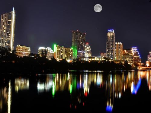 A vibrant city skyline at night, illuminated buildings reflecting in the water, with a full moon overhead.