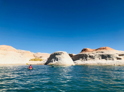 Two kayakers paddle in calm blue water near rocky cliffs under a clear blue sky.