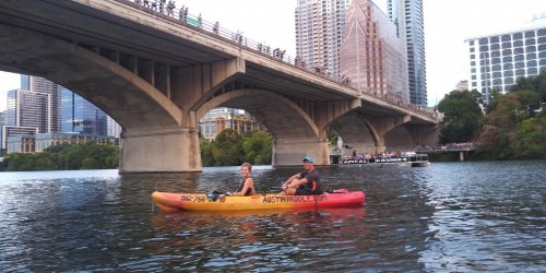 Two people in a yellow and red kayak on a river, with a city skyline and a bridge in the background.