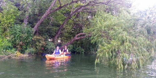 Two people in a kayak on a calm river, surrounded by lush greenery and overhanging trees.