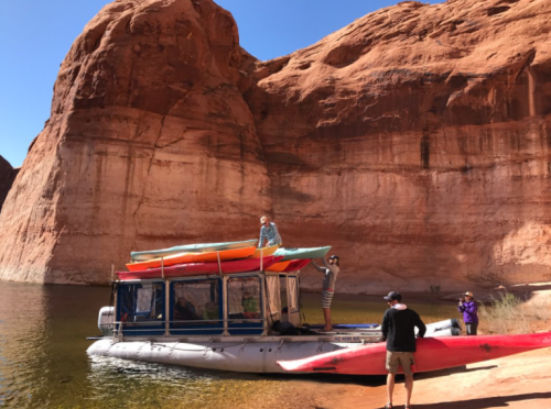 A group of people on a boat with kayaks on top, near a rocky canyon wall and calm water.