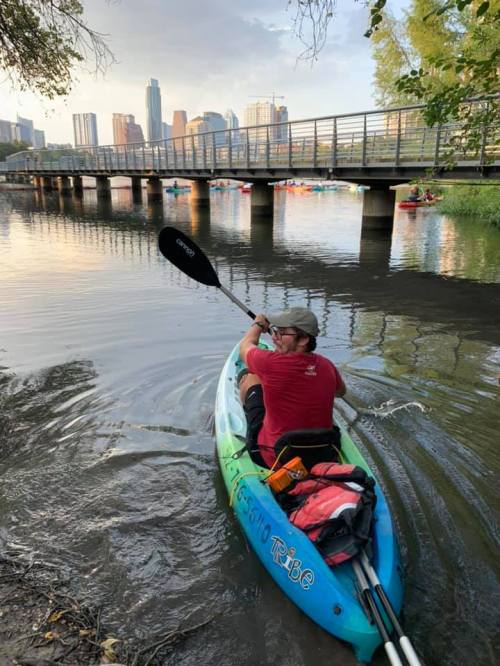 A person paddles a kayak on a calm river with a city skyline and a bridge in the background.
