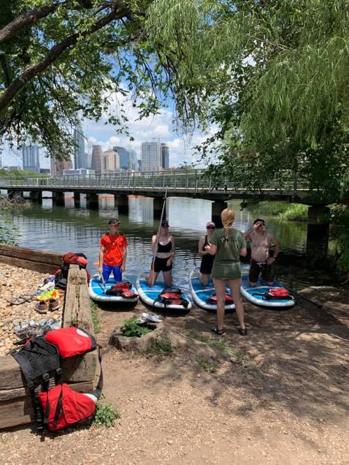 Group of people on paddleboards by a river, with a city skyline in the background and a bridge overhead.