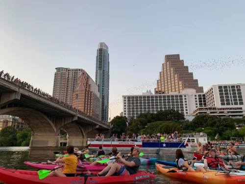 Kayakers on a river with a city skyline in the background, as a crowd watches from a bridge above.