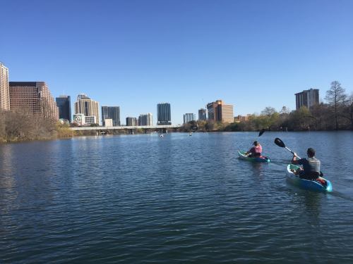 Two people kayak on a calm river with a city skyline in the background under a clear blue sky.