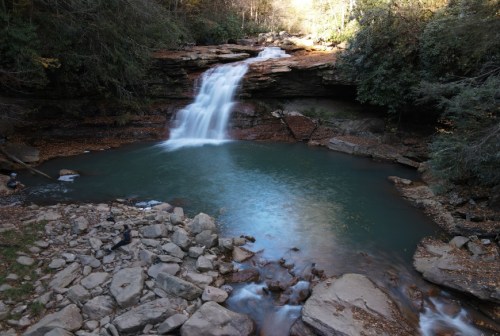 A serene waterfall cascades into a tranquil pool, surrounded by lush greenery and rocky terrain.