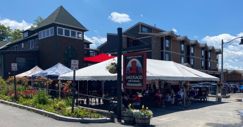 Outdoor seating area of Lake Placid Pub & Brewery, with a large tent and colorful flowers, against a sunny sky.