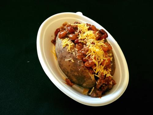 Baked potato topped with chili, beans, and shredded cheese, served in a white takeout container.
