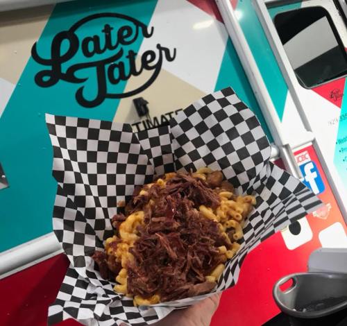 A hand holding a checkered paper bowl filled with macaroni and cheese topped with pulled pork, in front of a food truck.