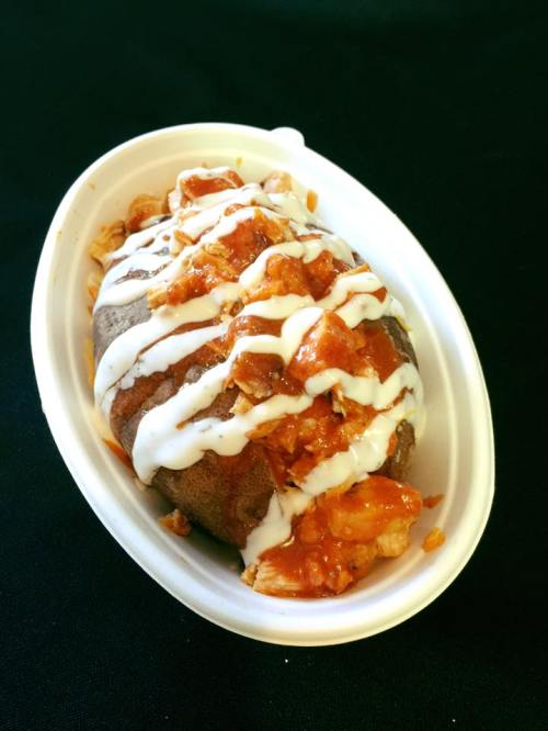 A baked potato topped with orange sauce, crumbled toppings, and drizzled with white sauce, served in a white container.