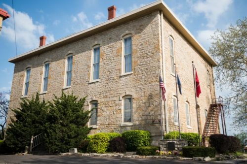 Historic stone building with flags, greenery, and a staircase, set against a blue sky with scattered clouds.