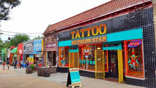 Colorful storefronts line a street, featuring a prominent tattoo shop with an "Open" sign and vibrant signage.