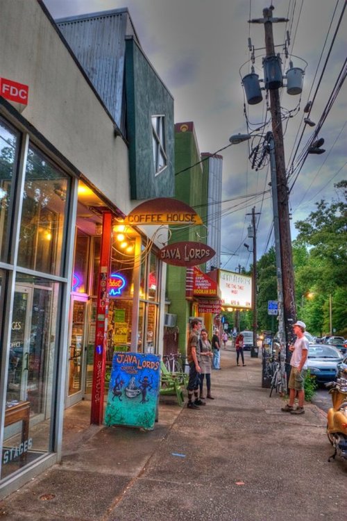A vibrant street scene featuring a coffee house, "Java Lords," with people and bicycles outside under a cloudy sky.