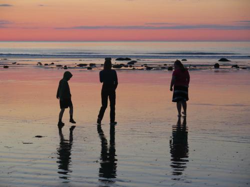 Three silhouetted figures walking along a beach at sunset, with colorful skies and reflections on the wet sand.