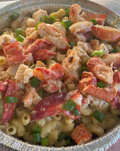 A close-up of macaroni topped with chunks of lobster and green onions in a foil container.