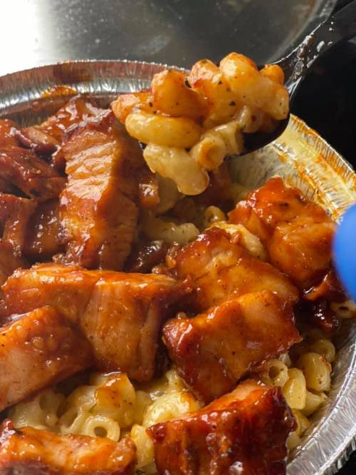 A close-up of a spoon lifting macaroni and cheese topped with pieces of glazed, grilled meat in a foil container.