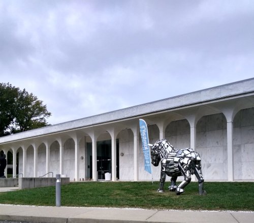 A metallic lion sculpture stands in front of a modern building with a blue banner, under a cloudy sky.