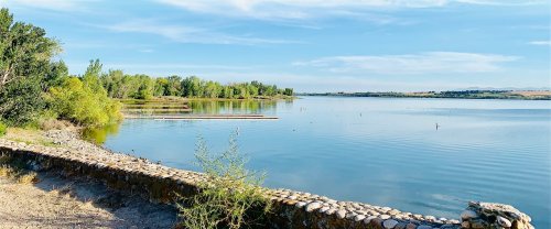 A serene lake surrounded by trees, with calm water reflecting the sky and a stone wall along the shore.