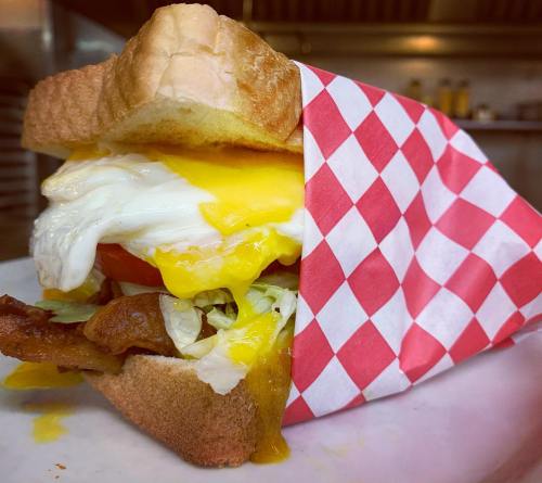 A close-up of a sandwich with bacon, lettuce, tomato, and a runny egg, wrapped in red and white checkered paper.