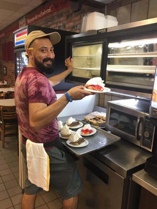 A man in a cap smiles while holding a tray of desserts near a microwave in a casual dining setting.