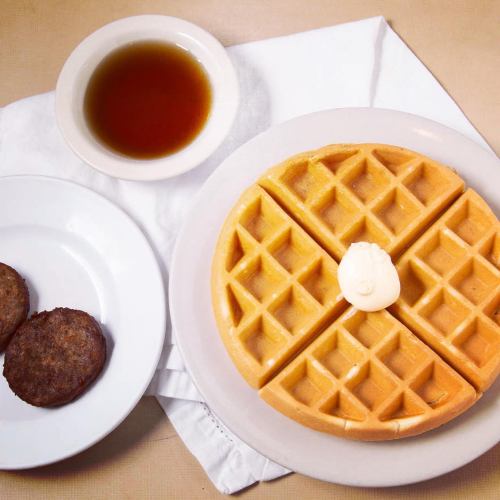 A plate with a golden waffle topped with butter, served with two sausage patties and a small bowl of syrup.