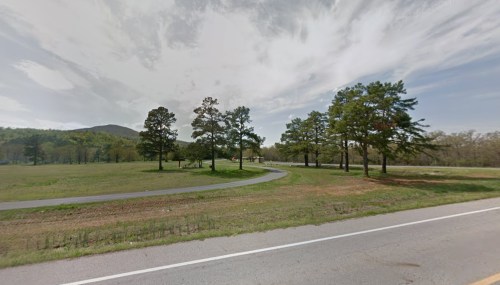 A winding road lined with trees, leading into a grassy area with distant hills under a cloudy sky.