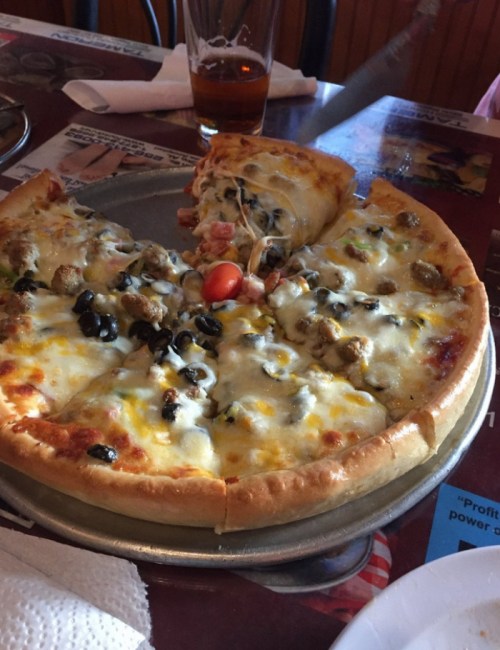 A freshly baked pizza with melted cheese, toppings including olives and tomatoes, served on a metal tray.