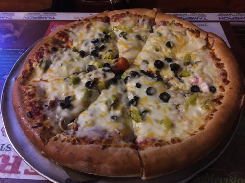 A large pizza topped with cheese, olives, peppers, and a cherry tomato, sliced into eight pieces on a metal tray.