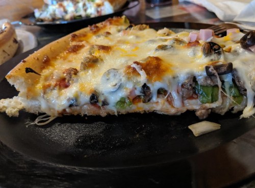 A close-up of a slice of pizza topped with cheese, olives, green peppers, and meat on a black plate.
