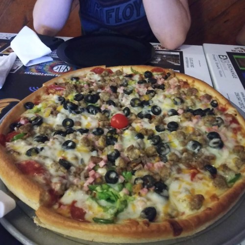 A large pizza topped with cheese, olives, ham, green peppers, and a cherry tomato in the center, on a metal tray.