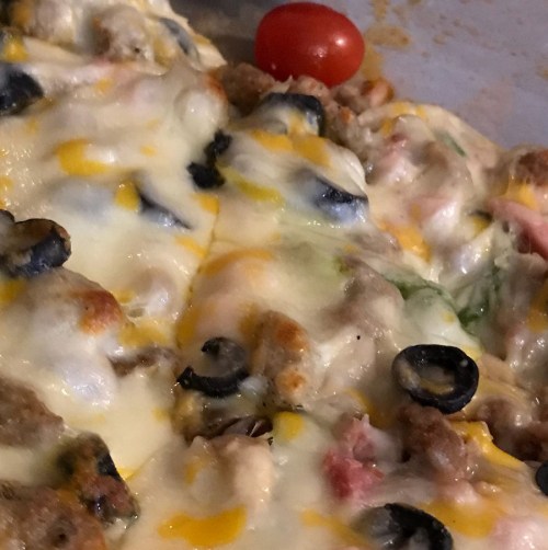 Close-up of a cheesy dish topped with olives, tomatoes, and ground meat, with melted cheese bubbling on top.