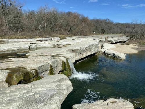 A rocky riverbank with a small waterfall, surrounded by trees and a clear blue sky.