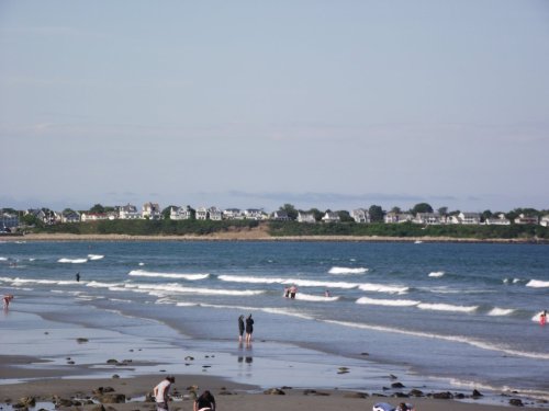 A beach scene with people walking along the shore, gentle waves, and a distant view of houses on a hillside.