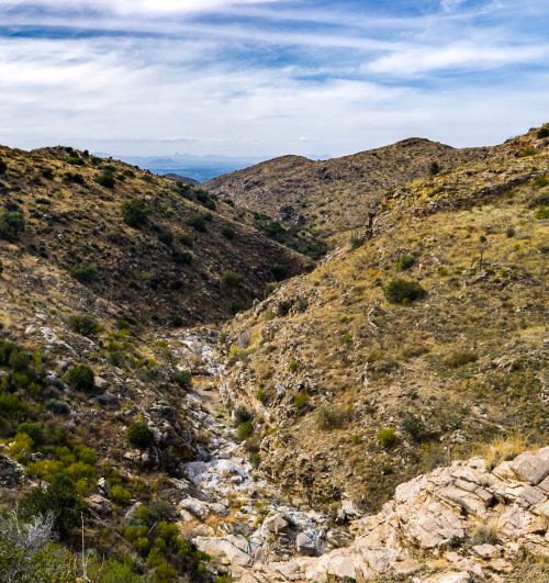 A scenic view of a rocky valley with sparse vegetation and distant hills under a partly cloudy sky.