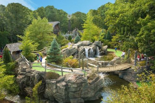 A scenic mini-golf course featuring a waterfall, bridge, and lush greenery, with people enjoying the outdoors.