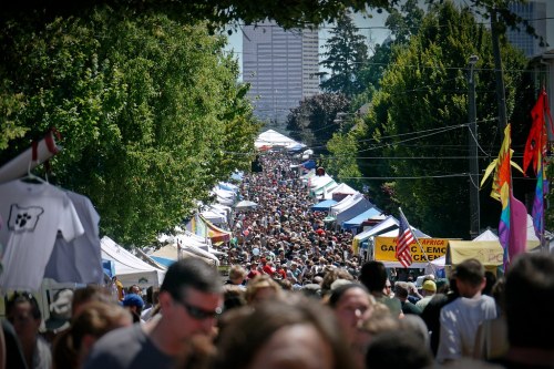 A bustling street fair filled with crowds, colorful tents, and trees lining the path under a clear blue sky.