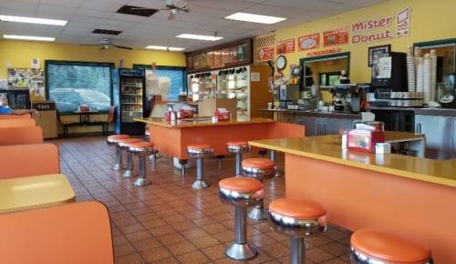 Interior of a donut shop with orange and silver decor, empty tables, and a counter displaying donuts and drinks.