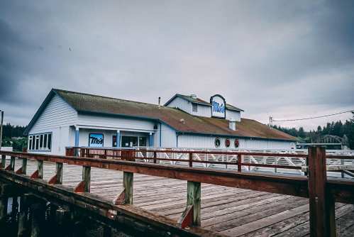 A blue building with a green roof, located on a pier, with a wooden walkway leading to it under a cloudy sky.