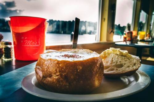 A bowl of clam chowder served in a bread bowl, with a piece of bread on the side, against a scenic window view.