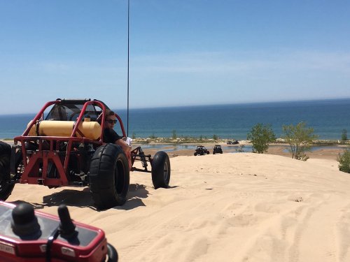 A red dune buggy on sandy terrain with a view of the ocean in the background and other buggies in the distance.