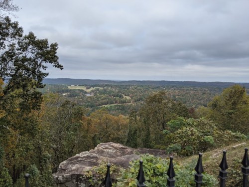 A scenic view from a rocky overlook, featuring rolling hills and trees in autumn colors under a cloudy sky.