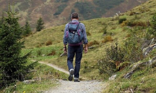 A person with a backpack walks along a gravel path through a green, hilly landscape.